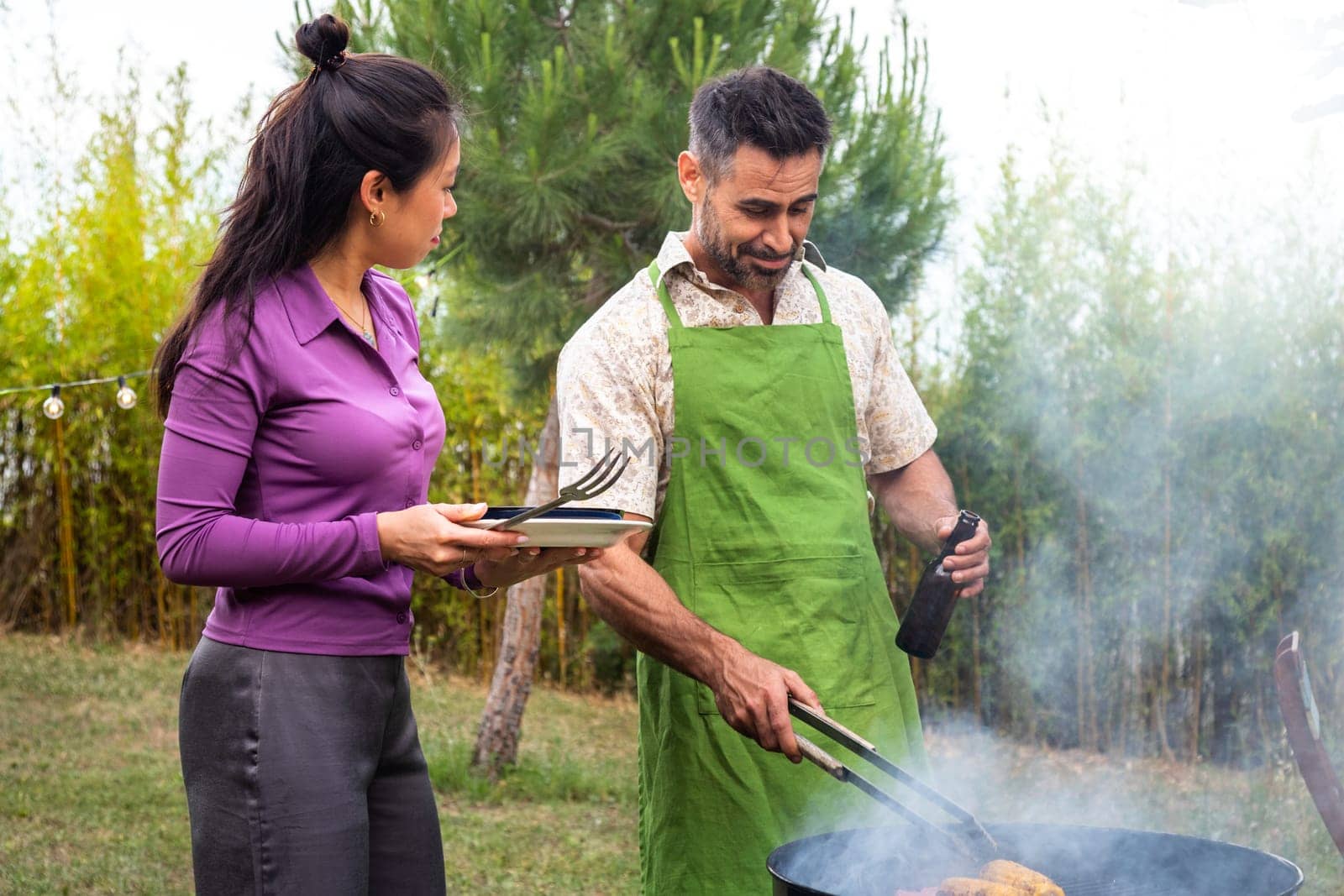 Asian young woman helping caucasian man cook some food on barbecue grill outdoors. Copy space. Garden dinner party concept.