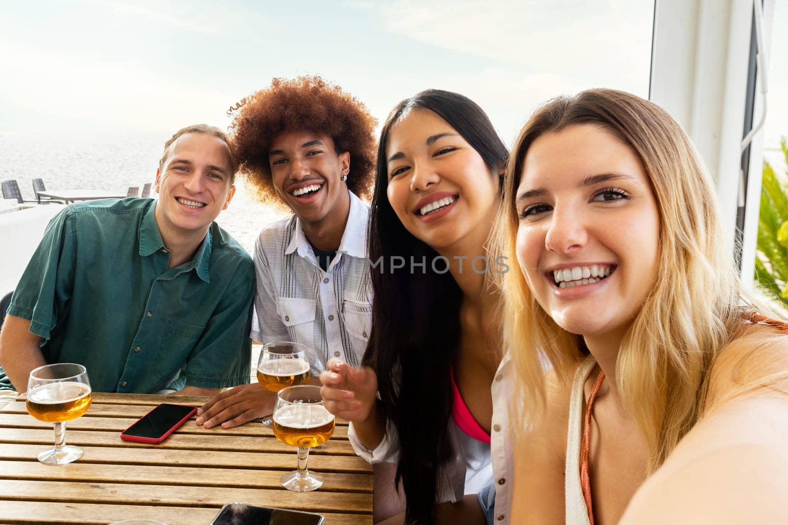 Multiracial group of friends taking selfie looking at camera while having drinks together at beach bar. Lifestyle and summertime concept.