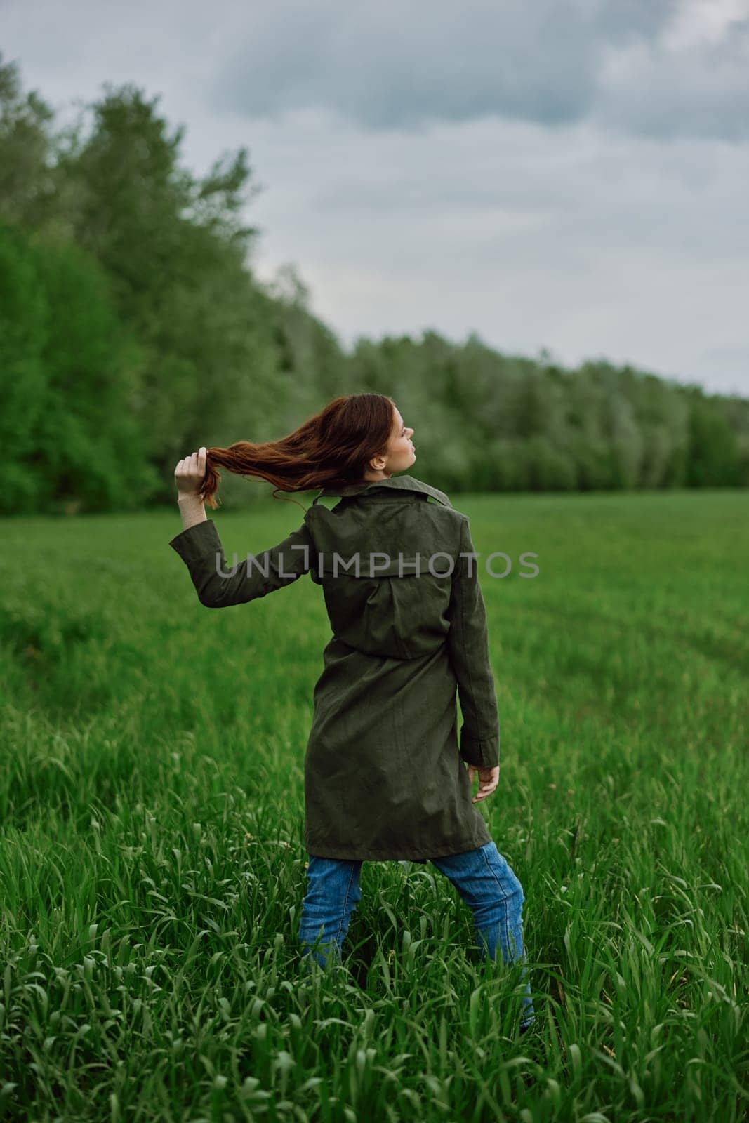 a beautiful woman in a dark coat stands in a green field in the spring in rainy weather and straightens her hair by Vichizh