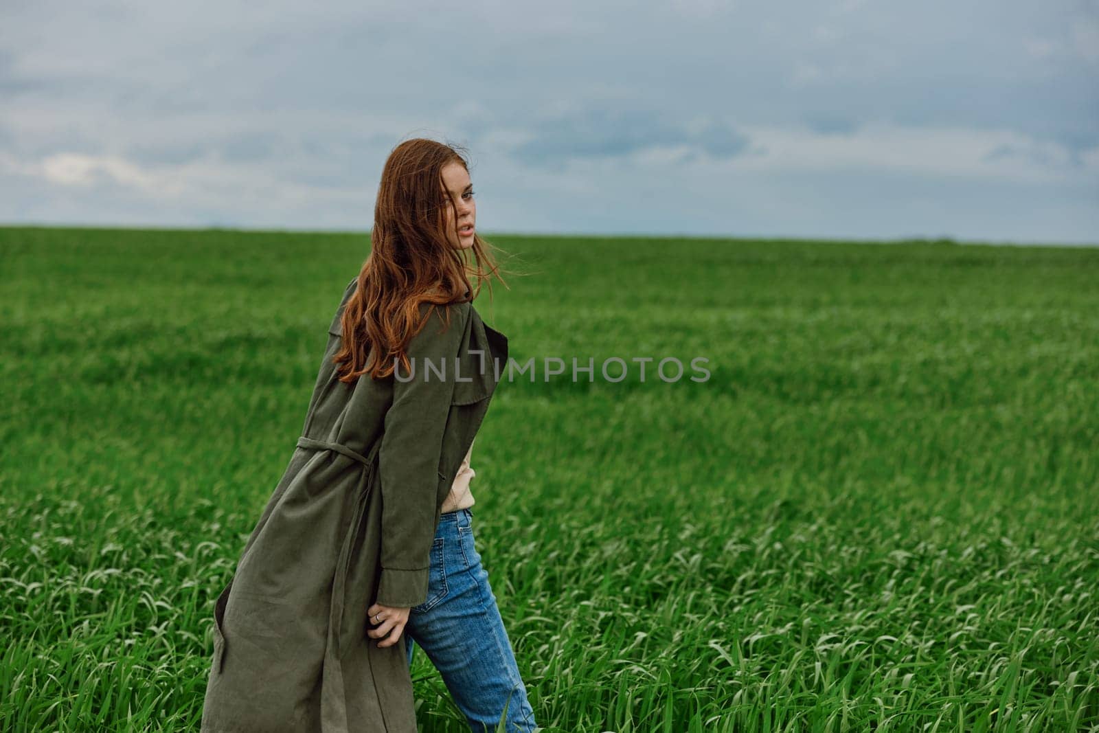 a woman in a dark coat stands in a green field in the wind close to the camera. Strong wind, cloudy weather. High quality photo