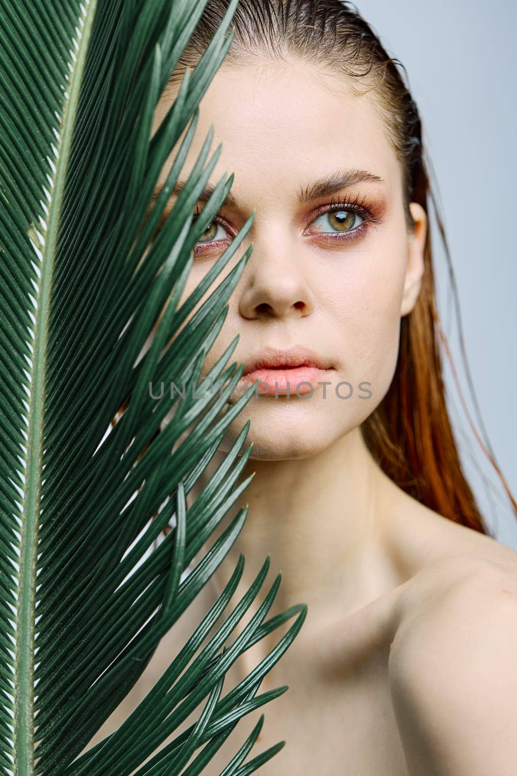 a close beauty portrait of a beautiful woman standing holding a tropical palm leaf in her hand, bringing it to her face. Vertical photo without retouching by Vichizh