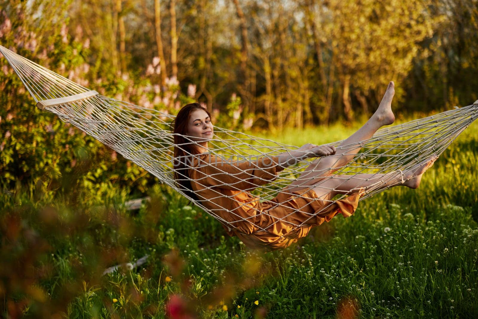 a joyful woman is sitting in a mesh hammock in nature relaxing and enjoying the rays of the setting sun looking into the camera, on a warm summer day. Horizontal photo on the theme of outdoor recreation. High quality photo