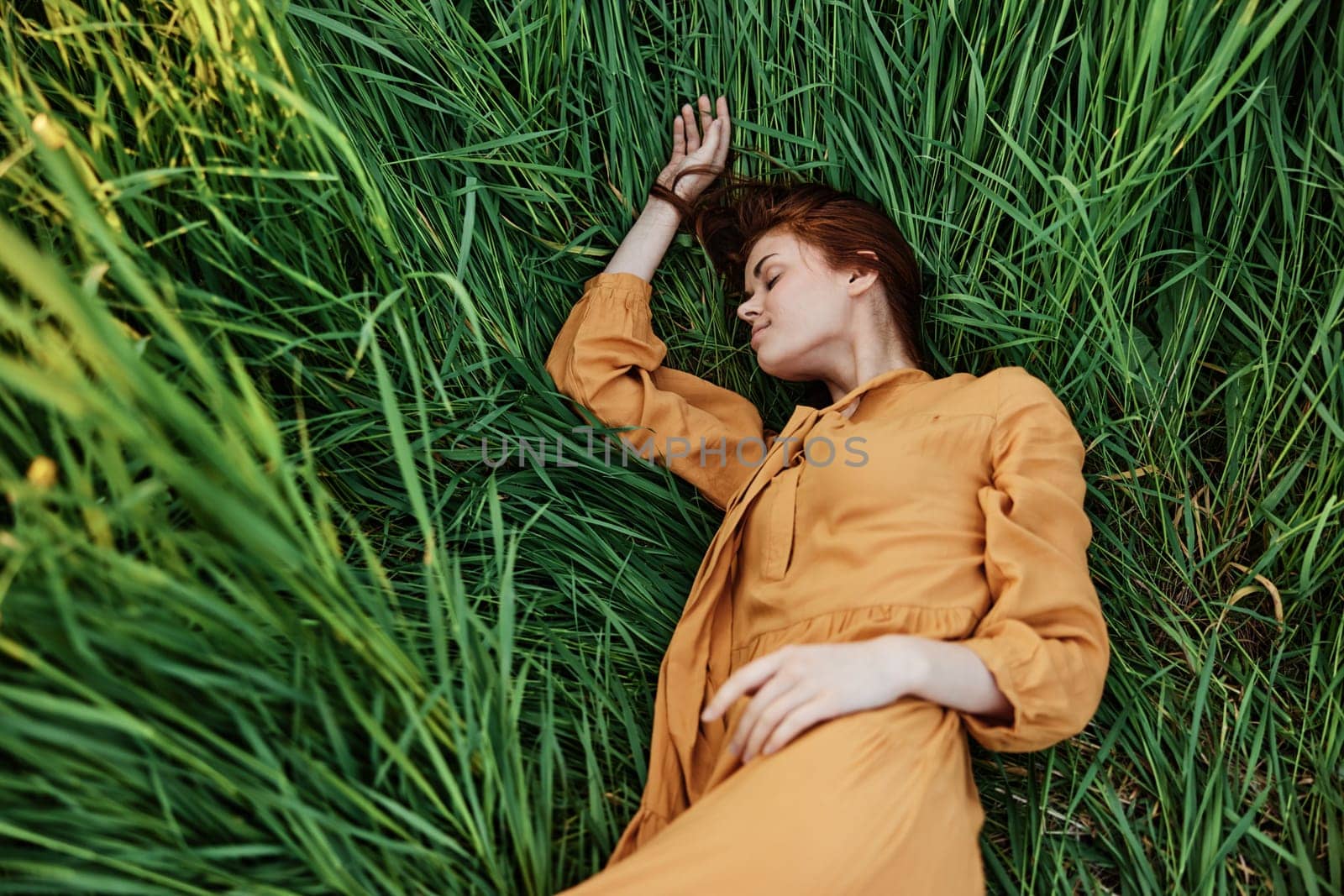 a close horizontal photo of a pleasant woman in a long orange dress resting lying in the tall grass with her eyes closed in sunny weather by Vichizh