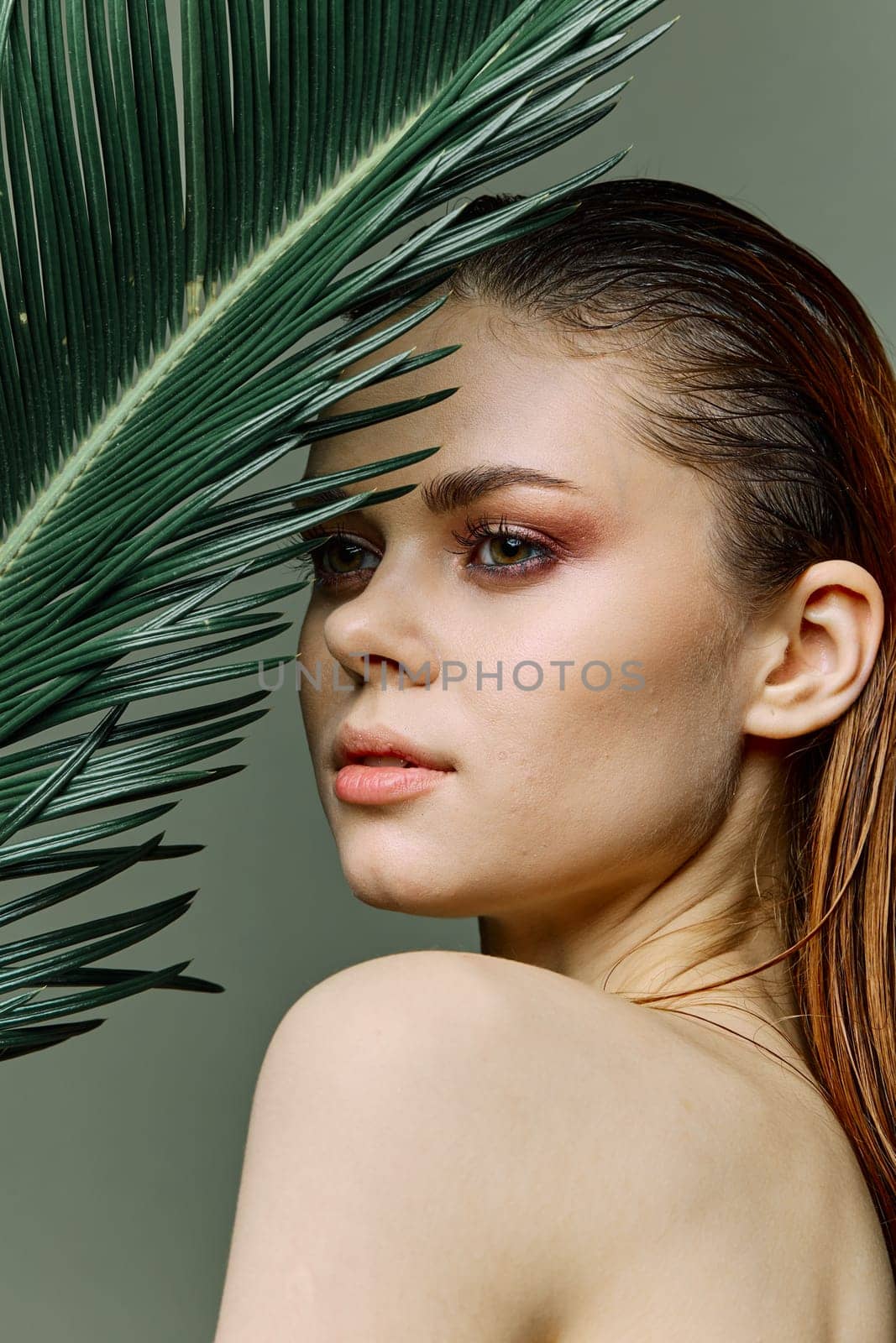 a sophisticated, elegant woman with evening makeup, stands with a green palm leaf, covering part of her face with it, looking away. Vertical photo without retouching. High quality photo