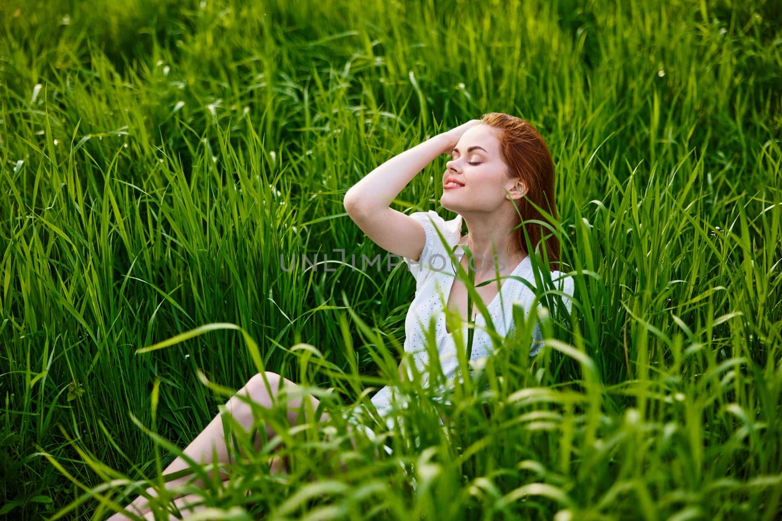 cute, happy woman sitting in tall grass on a sunny day in a light dress by Vichizh