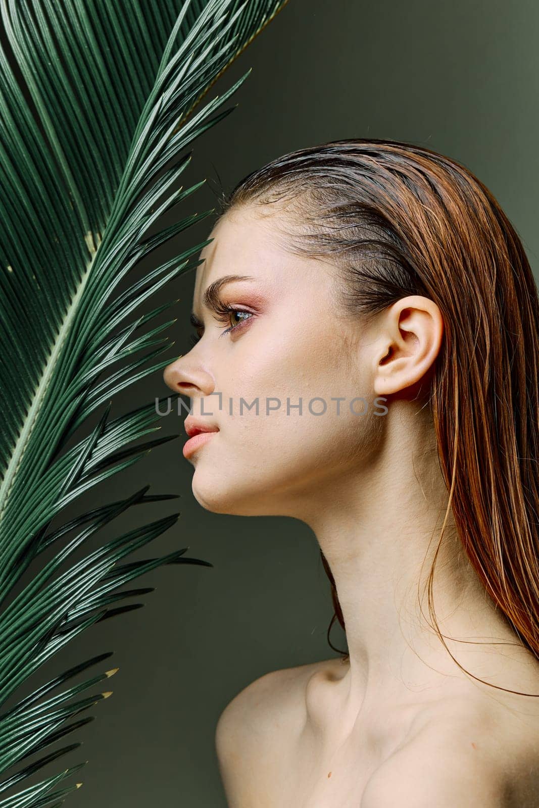 a sophisticated, elegant woman with evening makeup, stands with a green palm leaf, covering part of her face with it, looking away. Vertical photo without retouching by Vichizh
