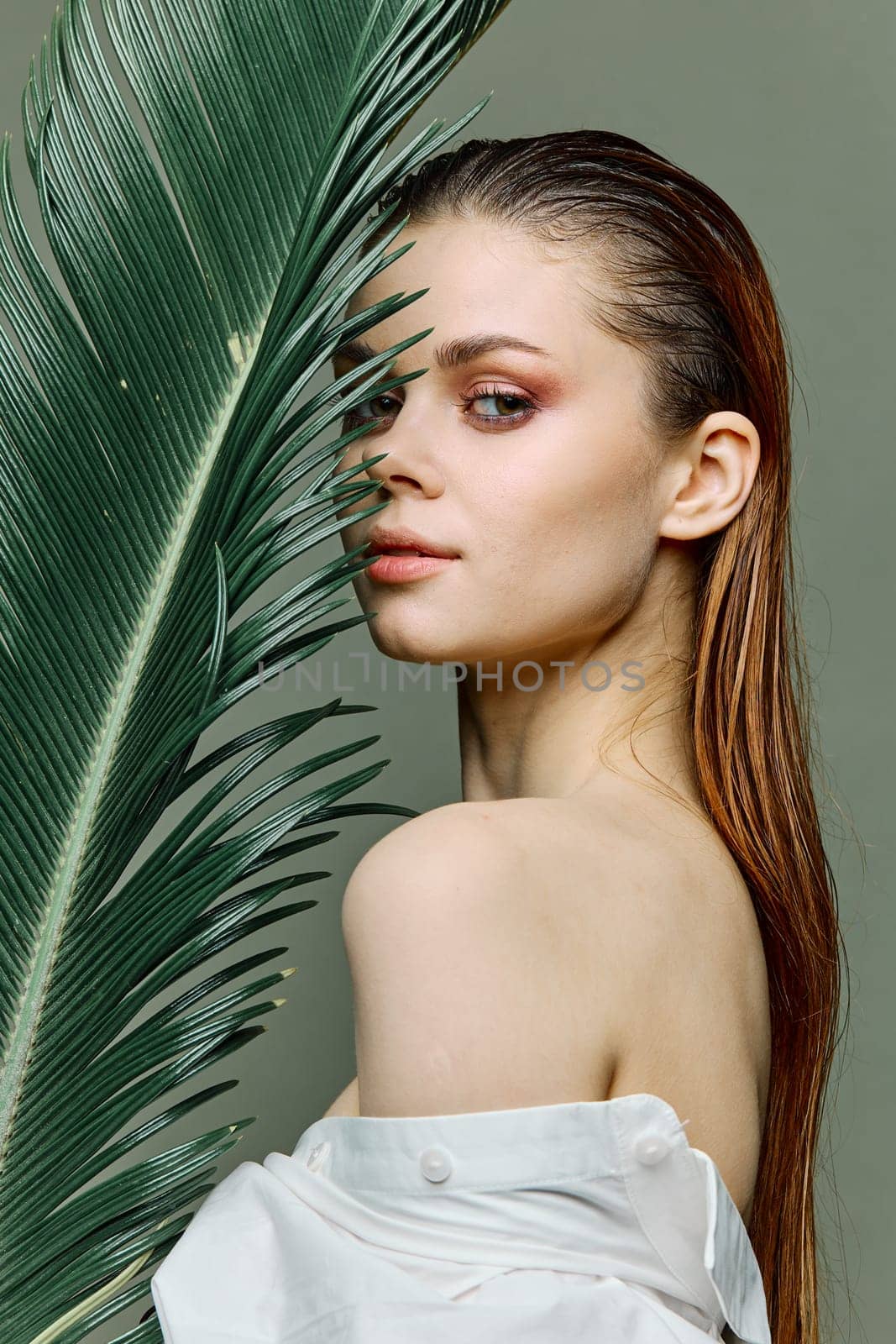 a sophisticated, elegant woman with evening makeup, stands with a green palm leaf, covering part of her face with it, dressed in a light shirt. Close vertical portrait, photo without retouching by Vichizh