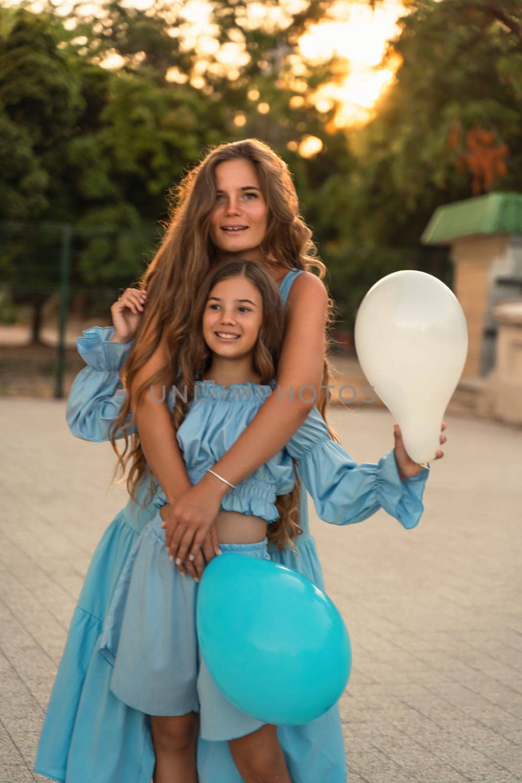 Mother daughter sunset. in blue dresses with flowing long hair against the backdrop of sunset. The woman hugs and presses the girl to her. They are looking at the camera. by Matiunina