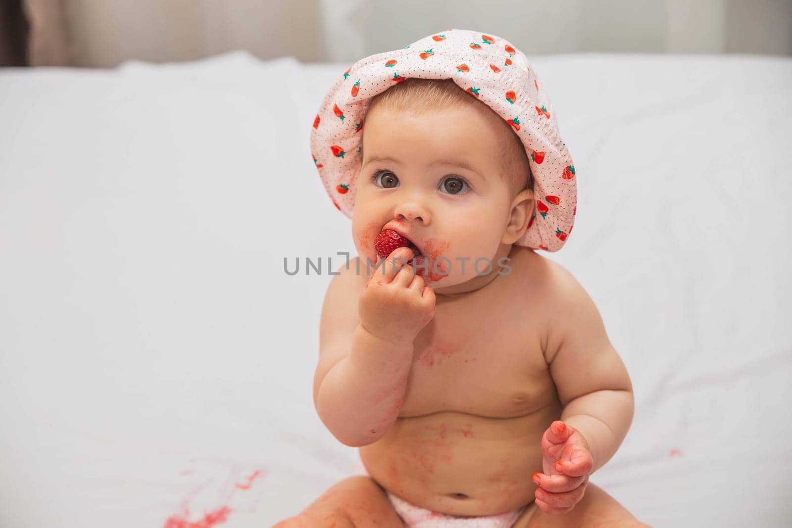 cute baby in a hat eating strawberries in bed.