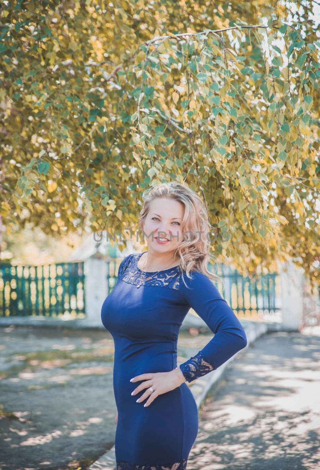 beautiful slim blonde with a long blue dress smiles under autumn tree.