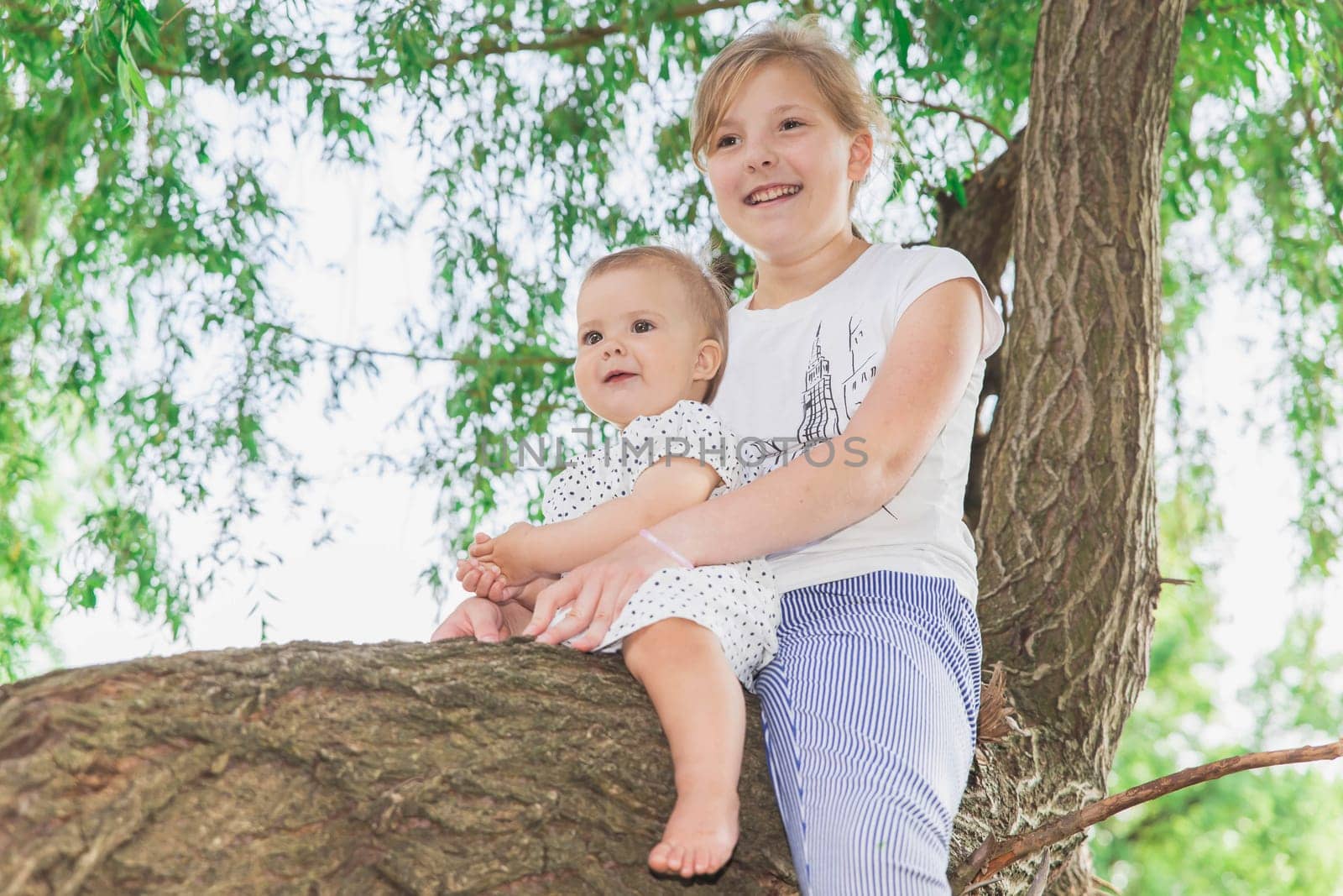 two smiling sisters are sitting on a tree and the older one is holding the younger