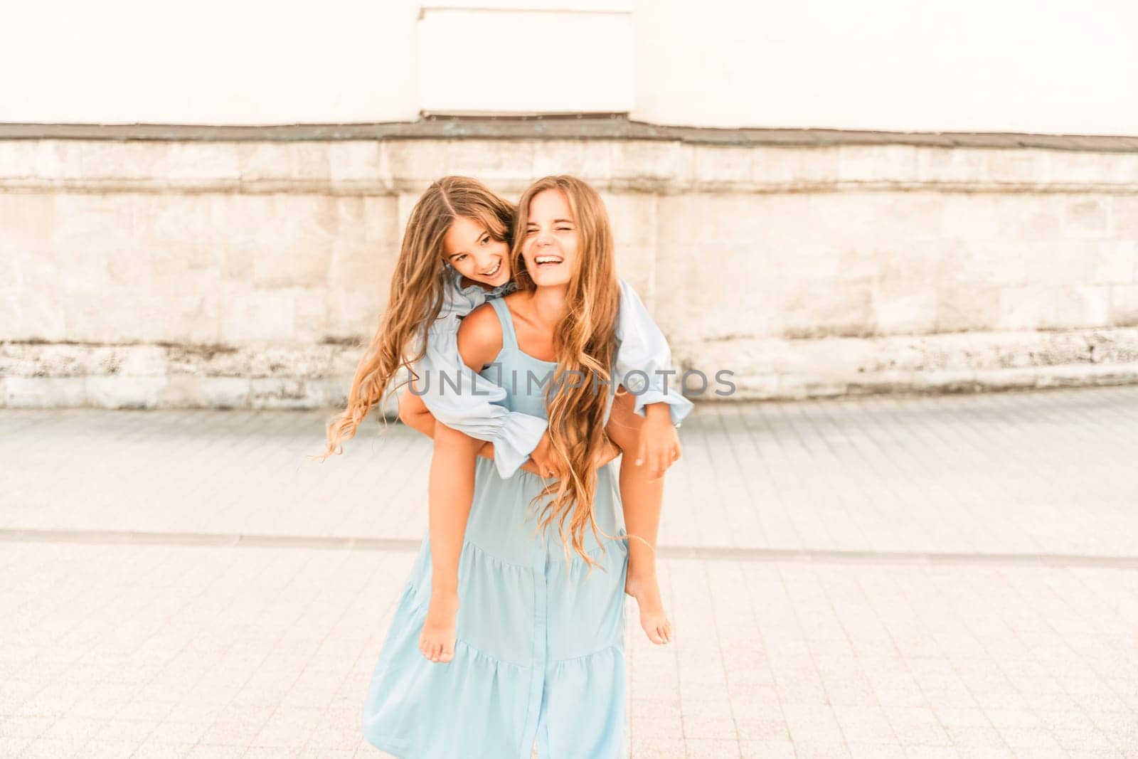 Mother of the daughter walks playing. Mother holds the girl on her back, holding her legs, and her daughter hugs her by the shoulders. Dressed in blue dresses. by Matiunina