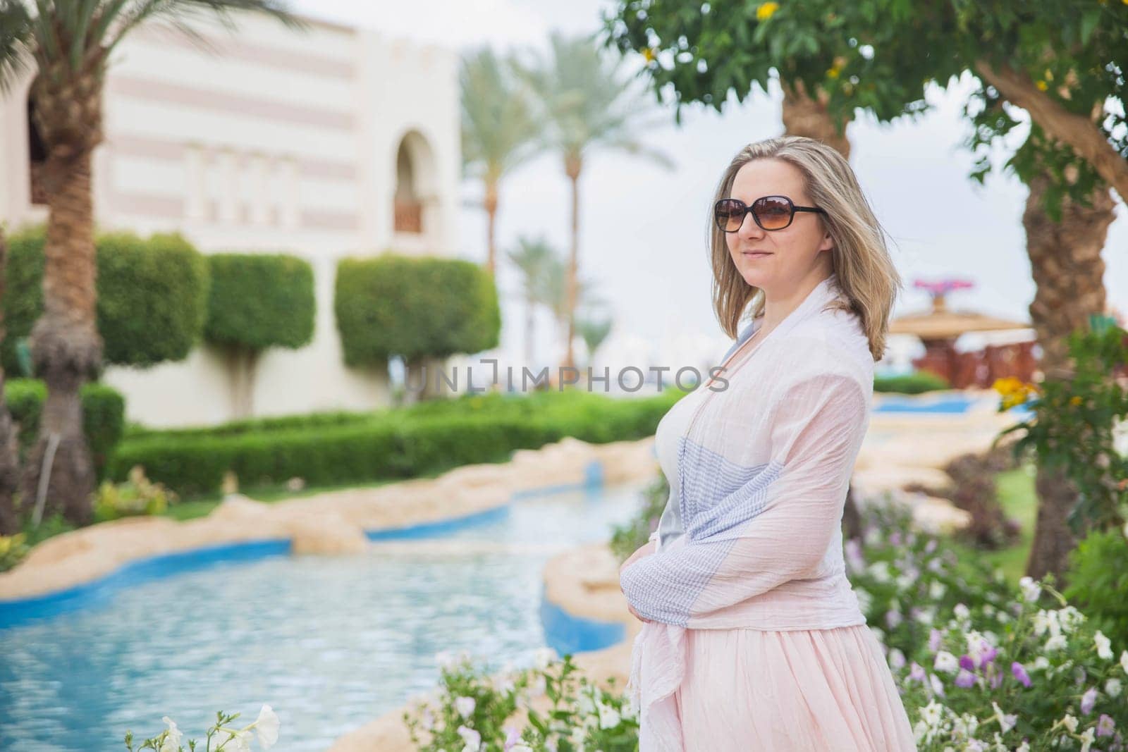 blonde in sunglasses and shawls standing in the garden with pool.