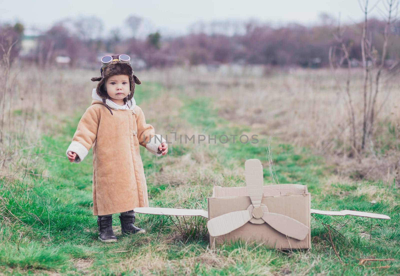 Charming baby in aviator's clothes stands near a cardboard plane.