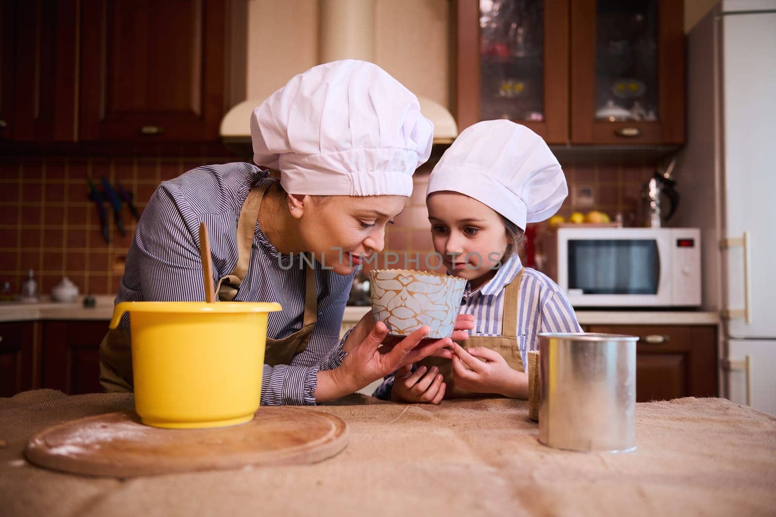 Happy family, loving mother and her little daughter, both of them wearing white chef's hat and beige apron, standing by kitchen island, sniffing dough while preparing cake together for Easter holiday