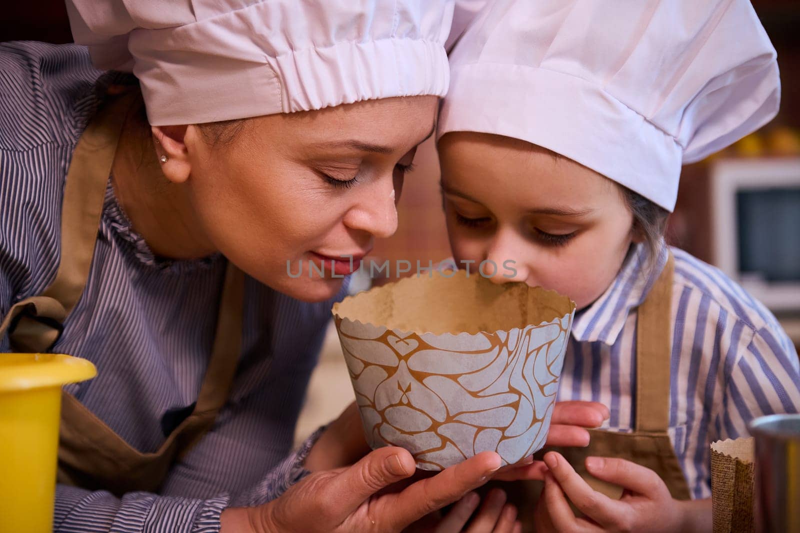 Close-up of a young multi-ethnic woman, a loving mother and her lovely little daughter, both of them wearing white chef's hat, sniffing dough while preparing Easter cake together in the home kitchen