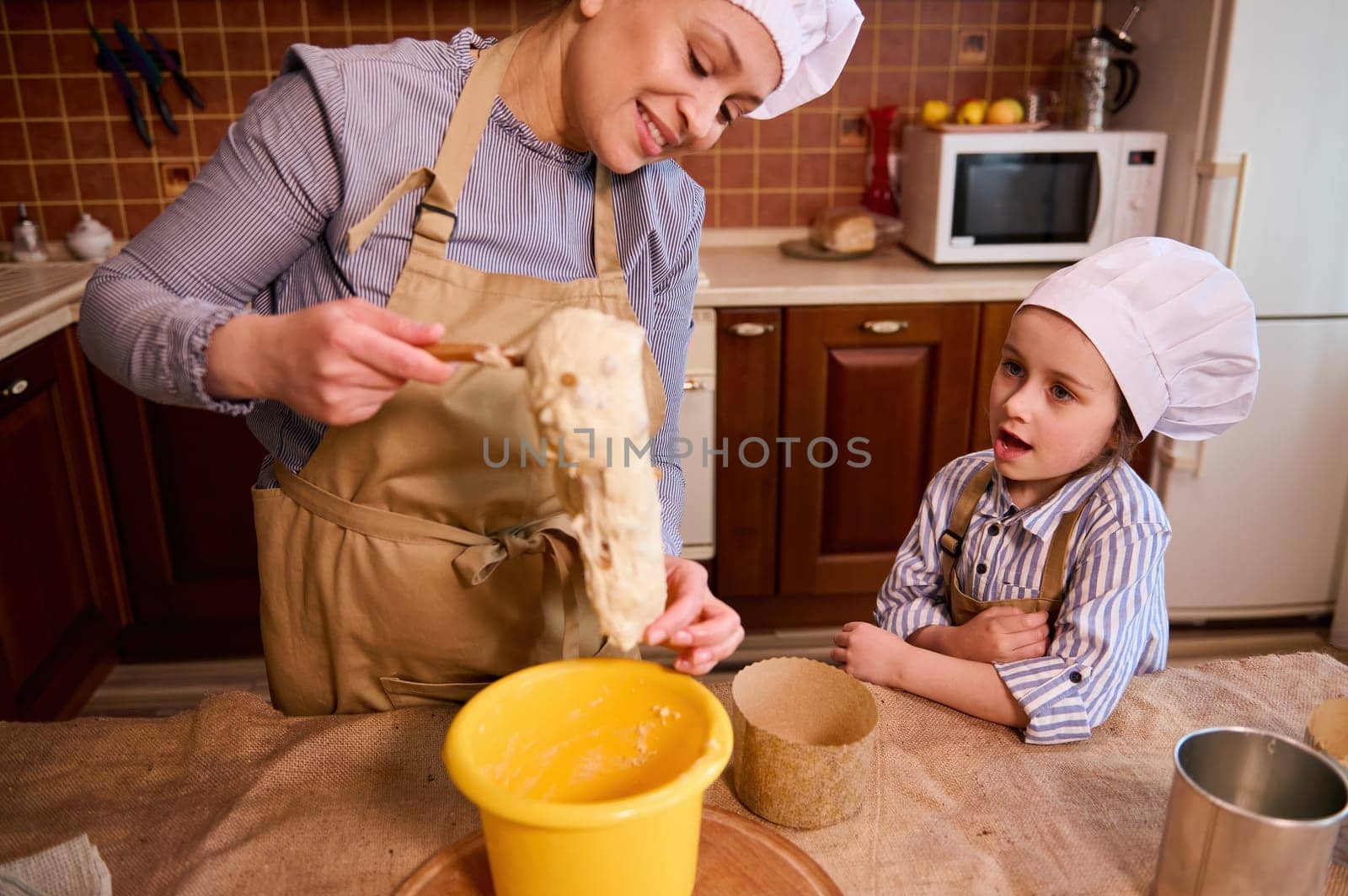 Amazed little toddler girl looking at her mom kneading dough, while preparing delicious homemade Easter cakes at home kitchen interior. Adorable daughter helping her mother preparing festive pastries