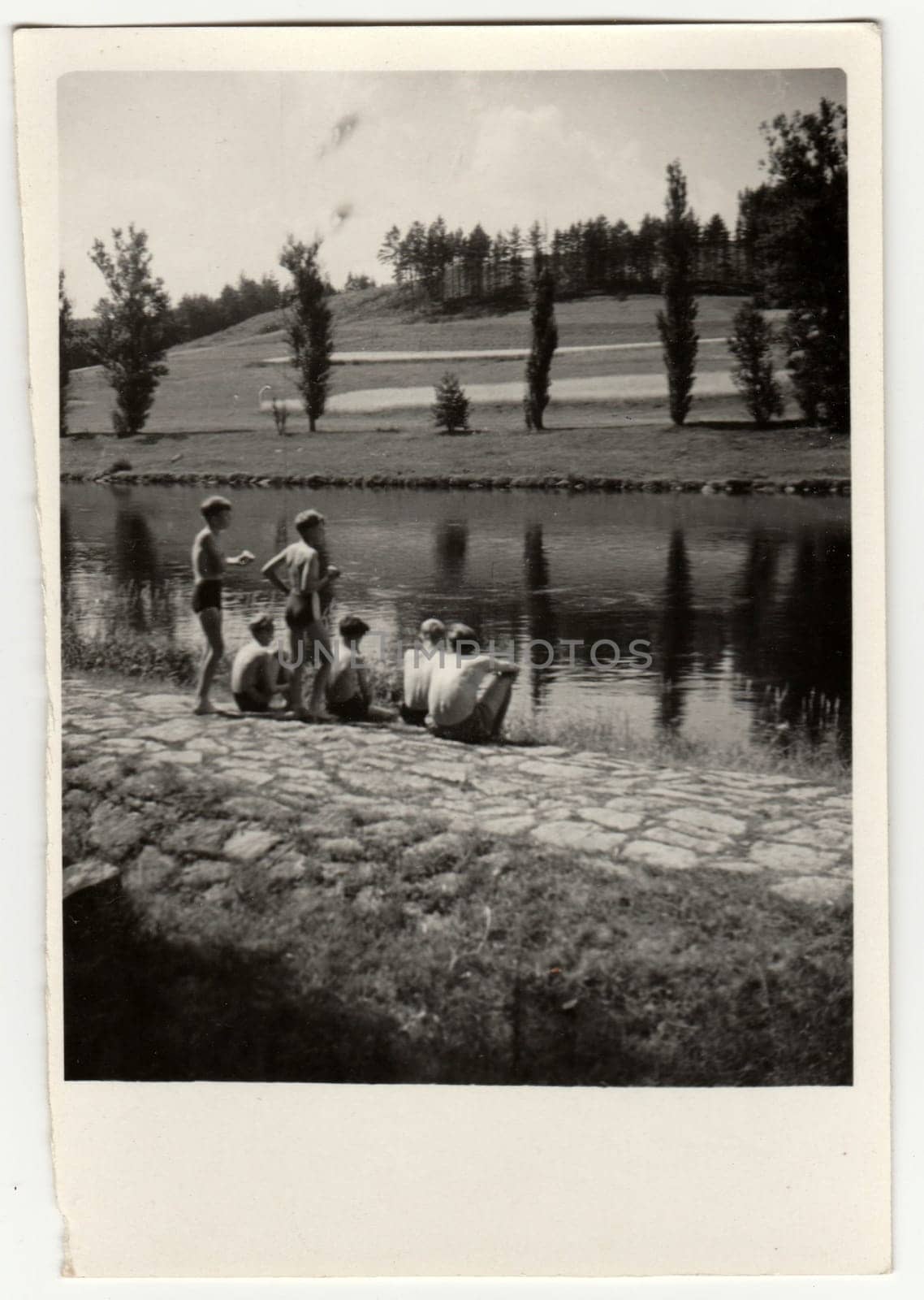 Retro photo shows a group of boys sit at the bank of river. Summer - vacation view. Vintage black and white photography. by roman_nerud