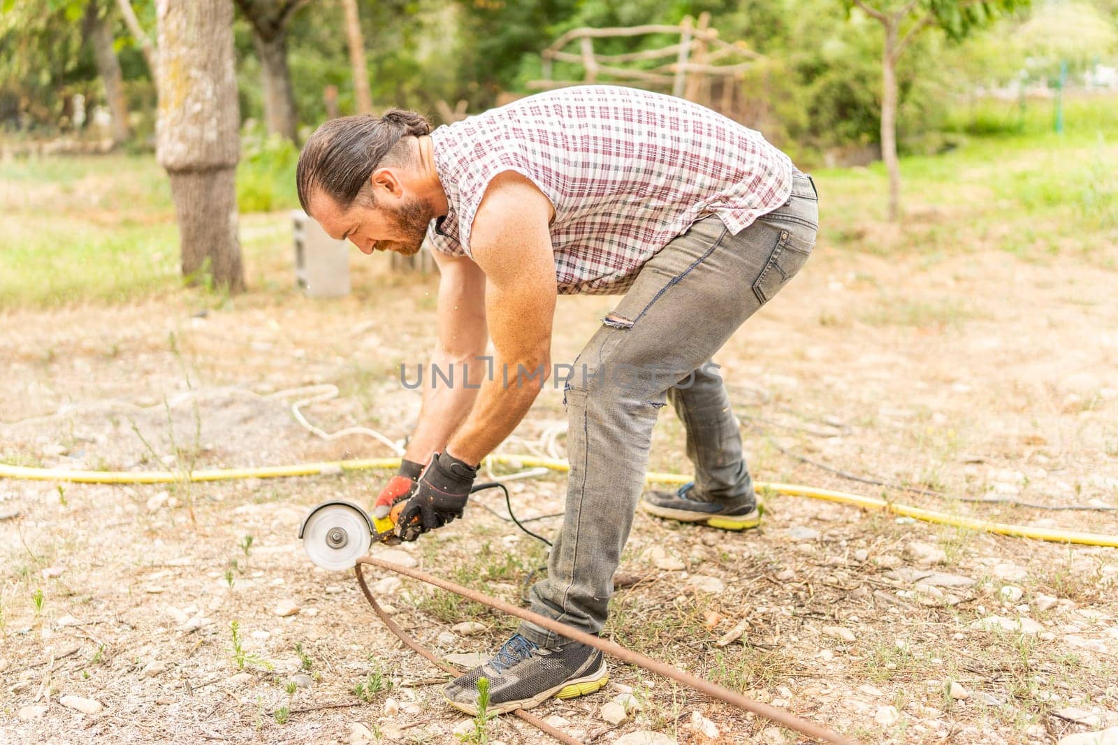 Man cutting a piece of iron using a radial saw in a yard with trees