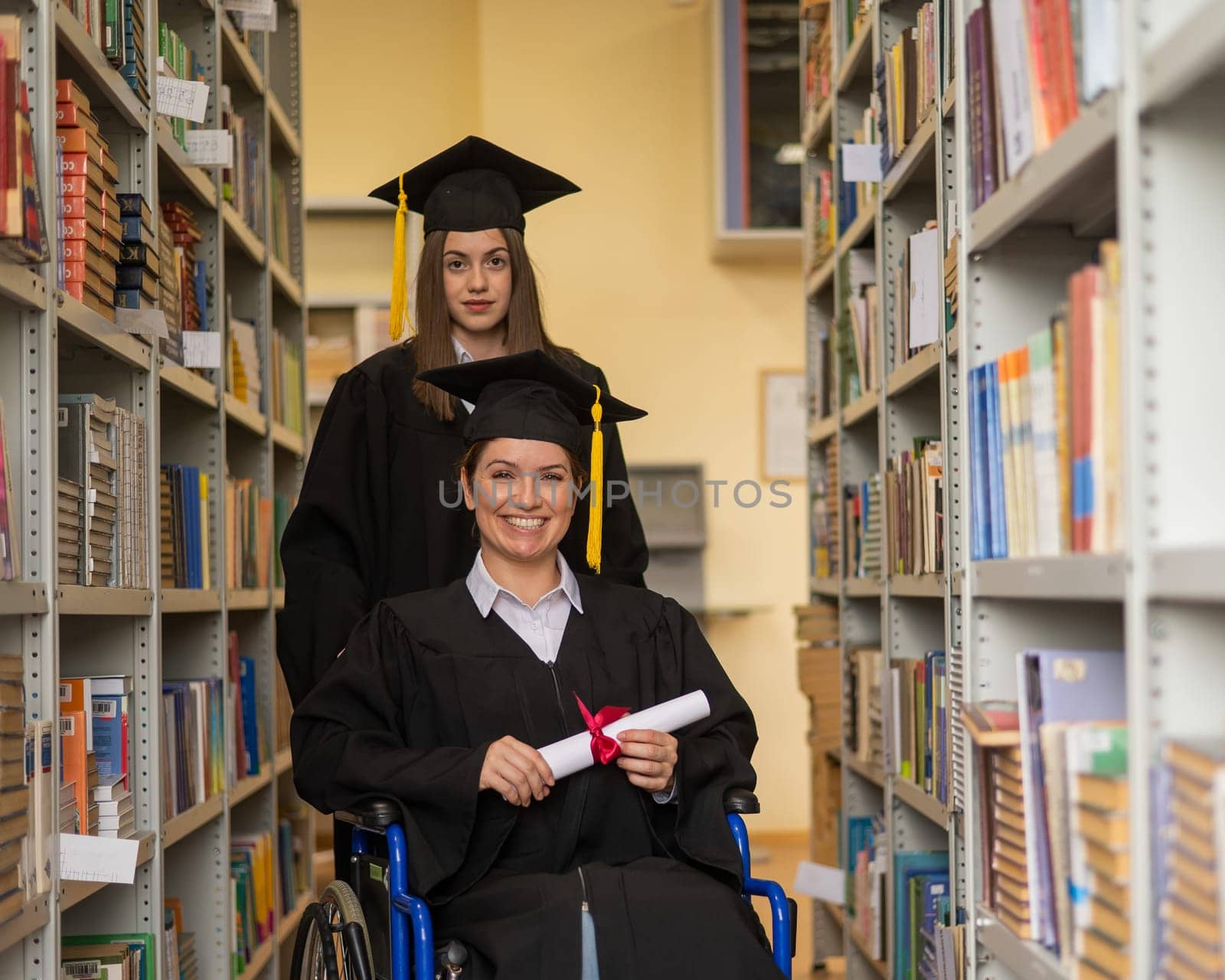 Happy young woman and woman in wheelchair in graduate gown with diploma in hands in library. Inclusive education. by mrwed54