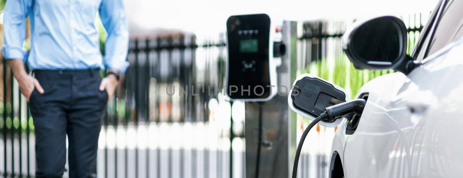 Closeup progressive suit-clad businessman with his electric vehicle recharge his car on public charging station in modern city with power cable plug and renewable energy-powered electric vehicle.