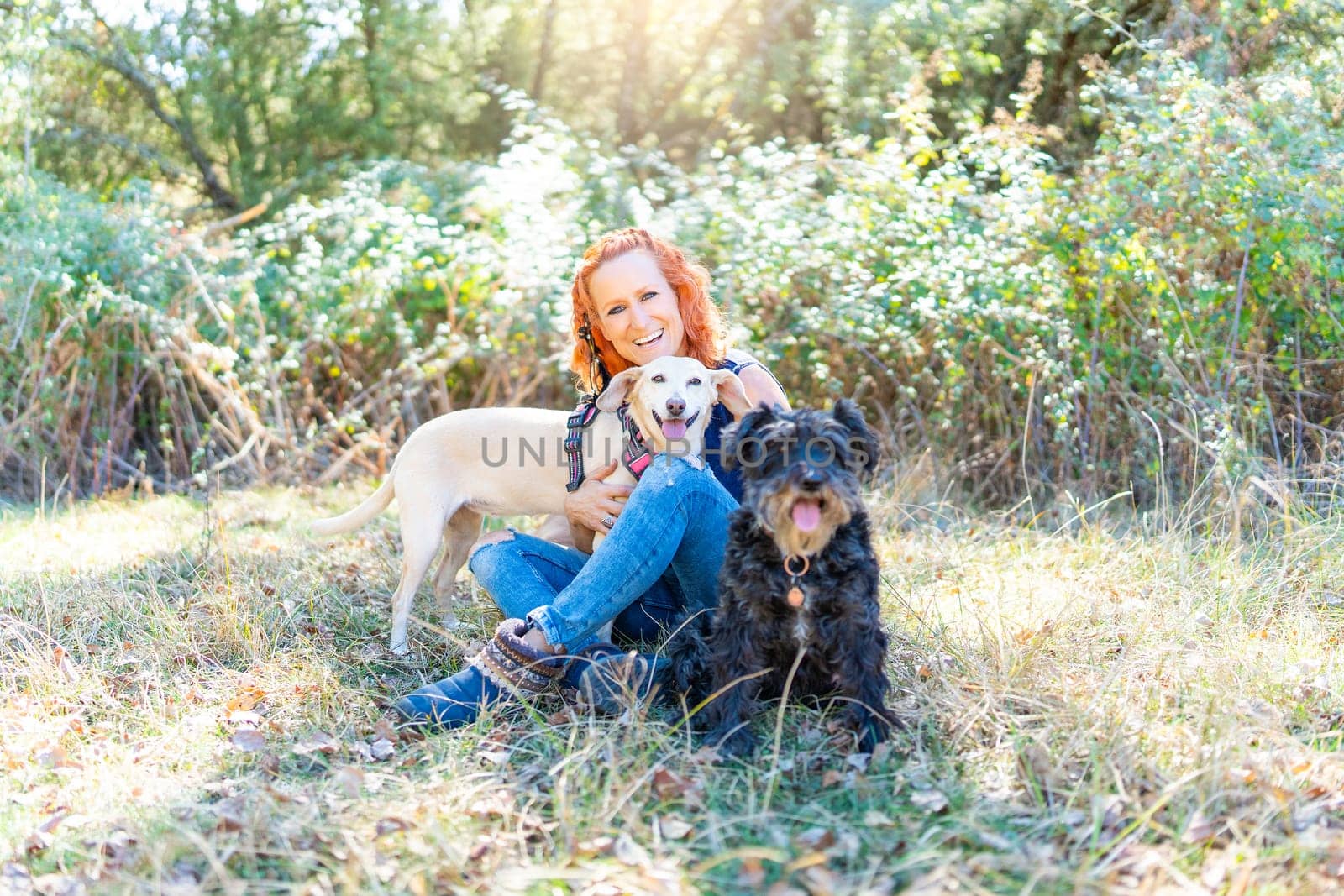 Portrait of a caucasian happy woman looking at the camera while embracing two dogs sitting in a field