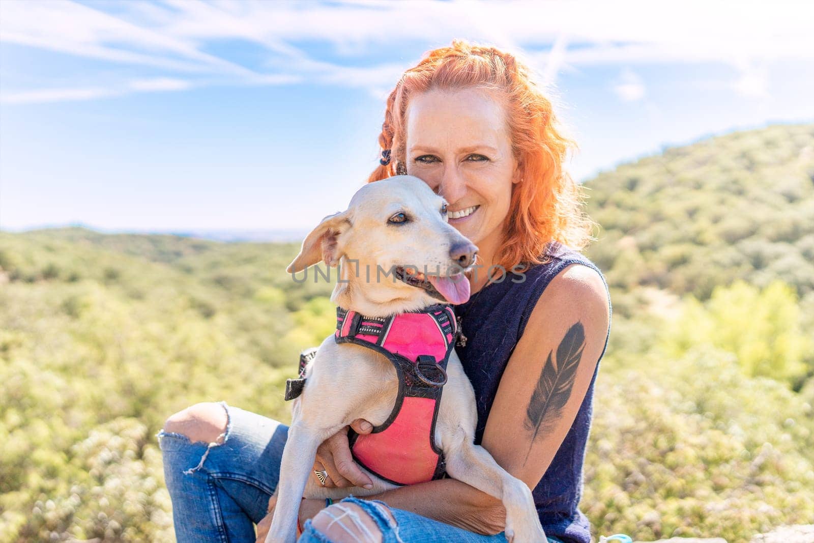 Caucasian woman smiling at the camera while embracing a white dog while sitting in the mountain