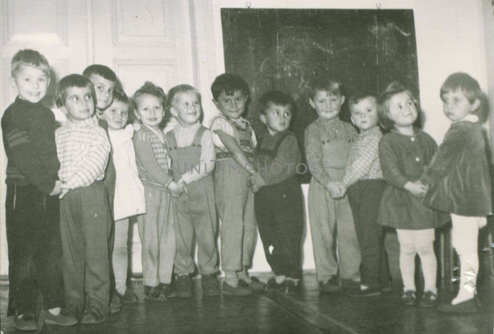 Retro photo shows small pupils in the classroom. Blackboard on the background. Vintage black and white photography. by roman_nerud