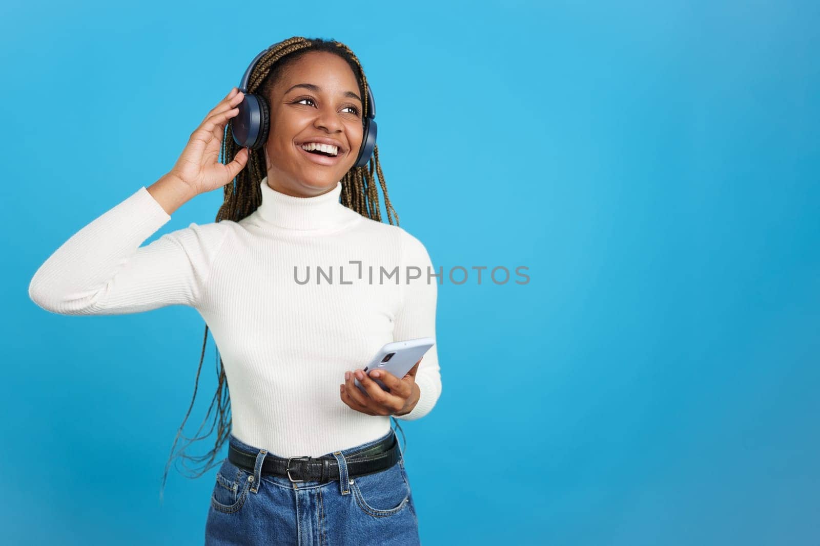 Studio portrait with blue background of a cool african woman listening to music with headphones and mobile