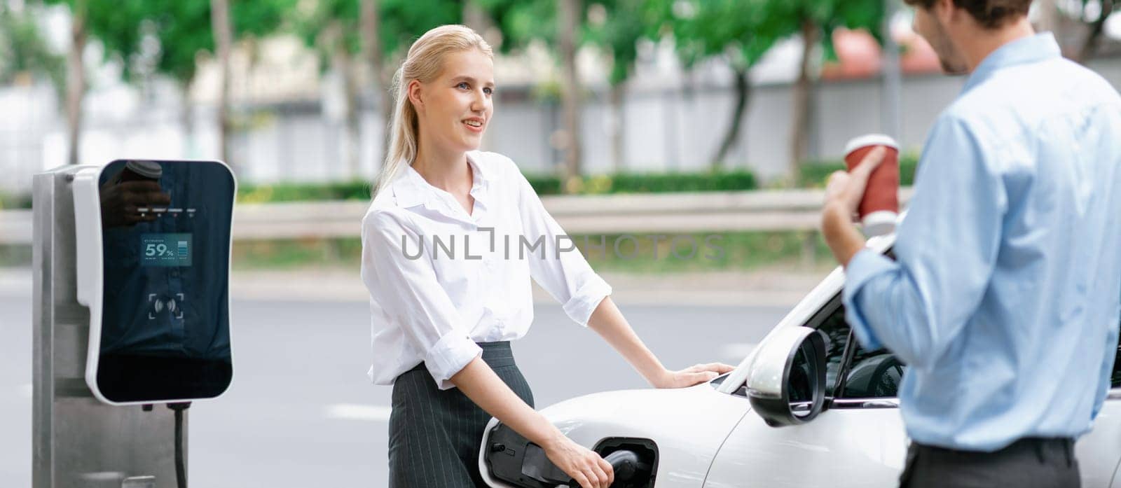 Progressive businessman and businesswoman with electric car parking and connected to public charging station before driving around city center. Eco friendly rechargeable car powered by clean energy.