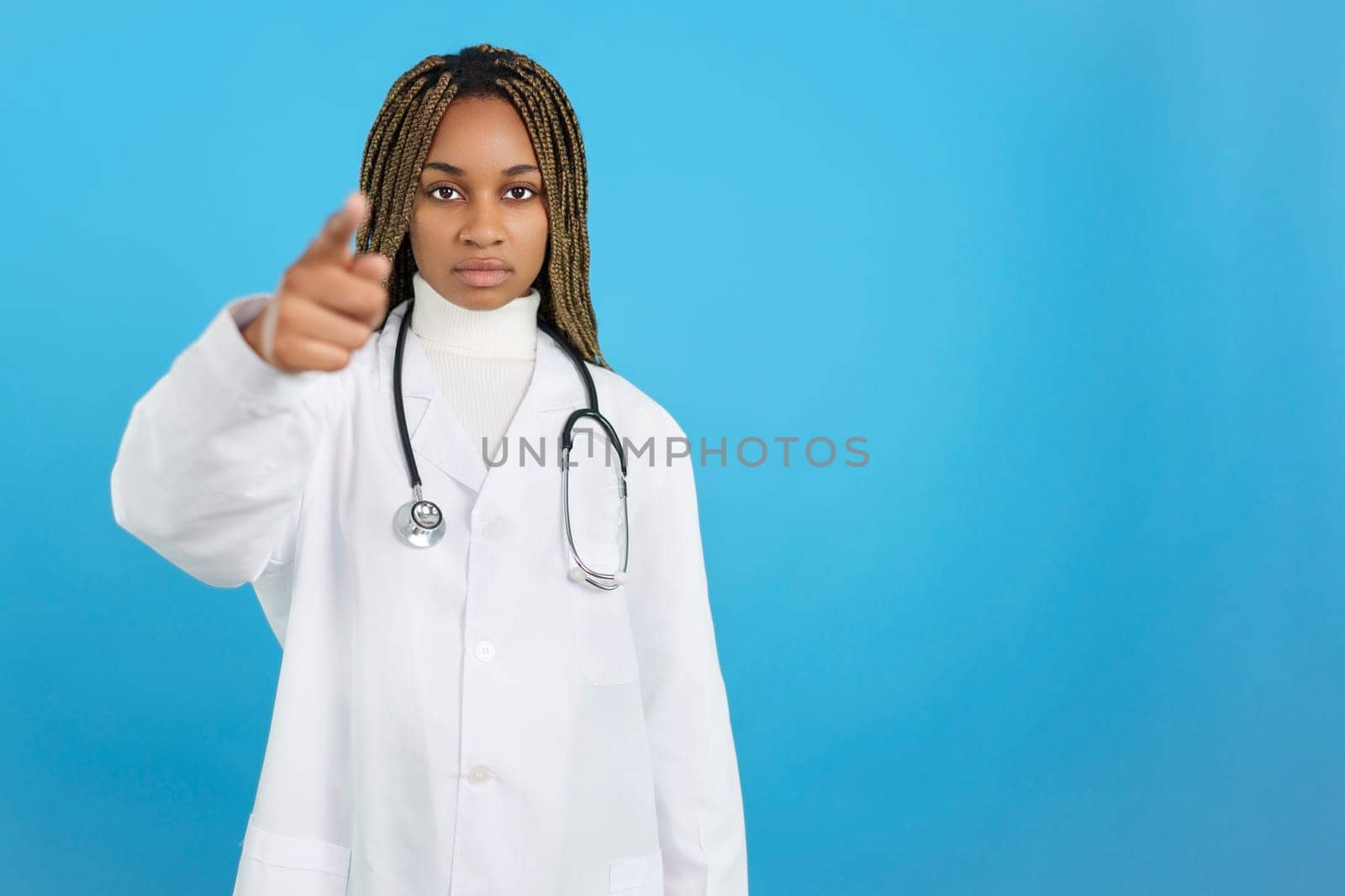 Studio portrait with blue background of a serious female african doctor wearing uniform pointing forward