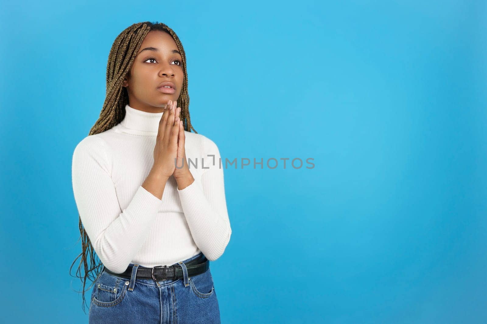 Studio portrait with blue background of a serious african woman looking up while praying with the hands folded