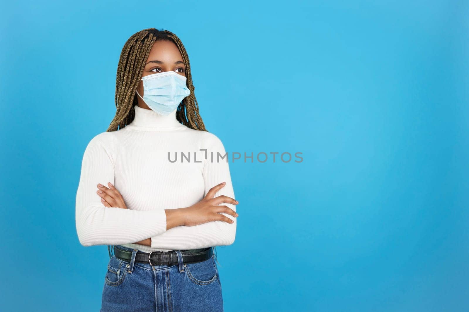 Studio portrait with blue background of a african woman standing with arms crossed wearing a facial protective mask