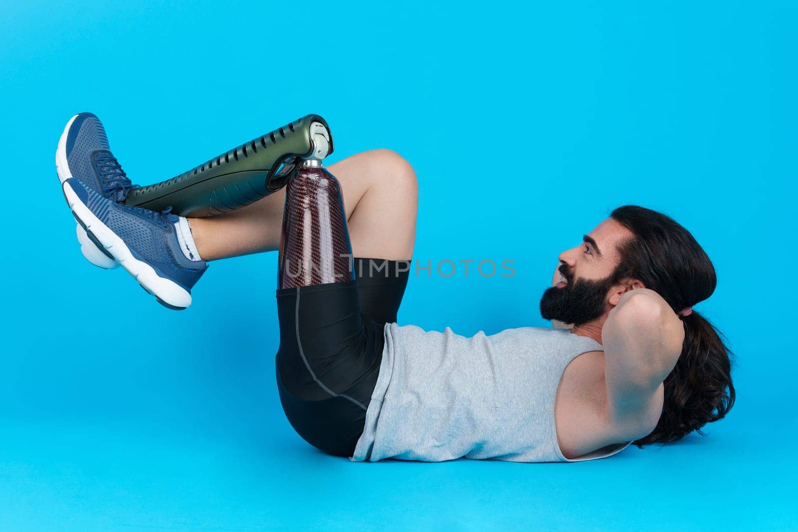 Horizontal studio portrait with blue background of a sportive bearded man with a prosthetic leg doing sit-ups