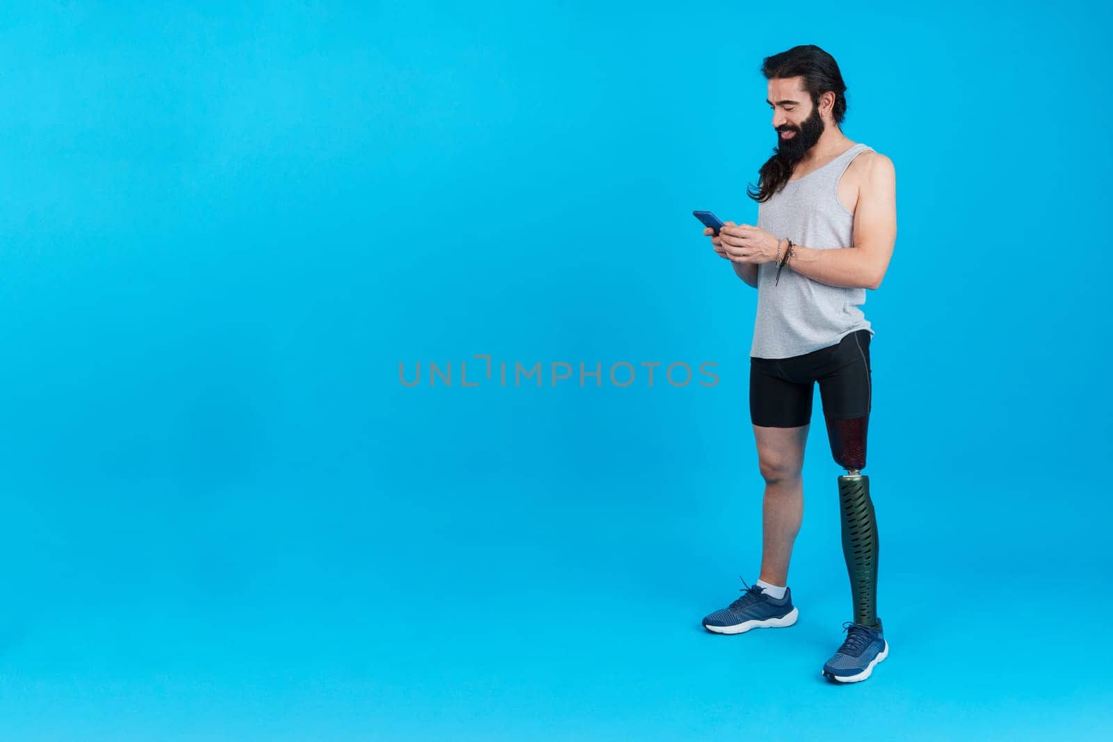 Studio portrait with blue background and copy space of a sportive man with a leg prosthesis using a mobile