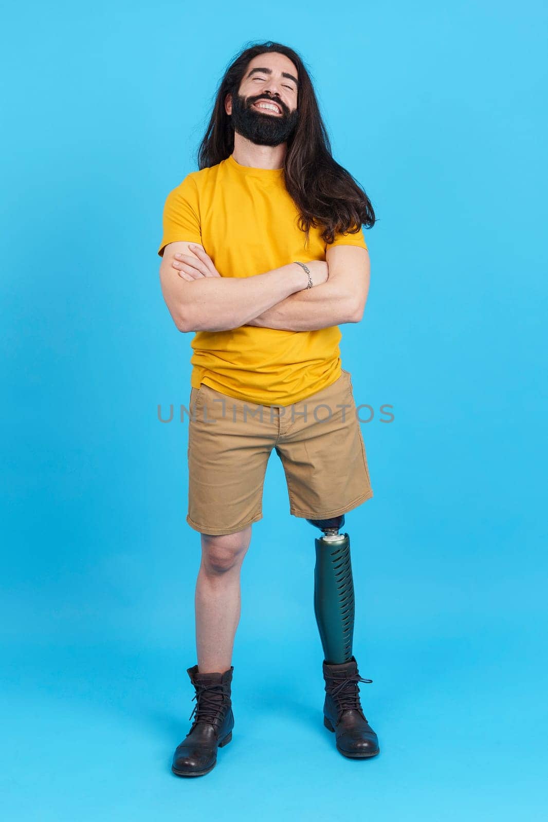 Vertical studio portrait with blue background of a cool man with a leg prosthesis standing with arms crossed