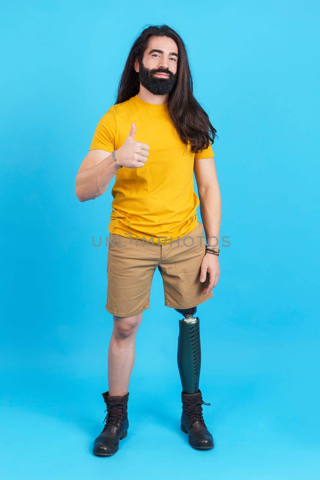 Vertical studio portrait with blue background of a man with a prosthesis leg gesturing approval with the thumb up
