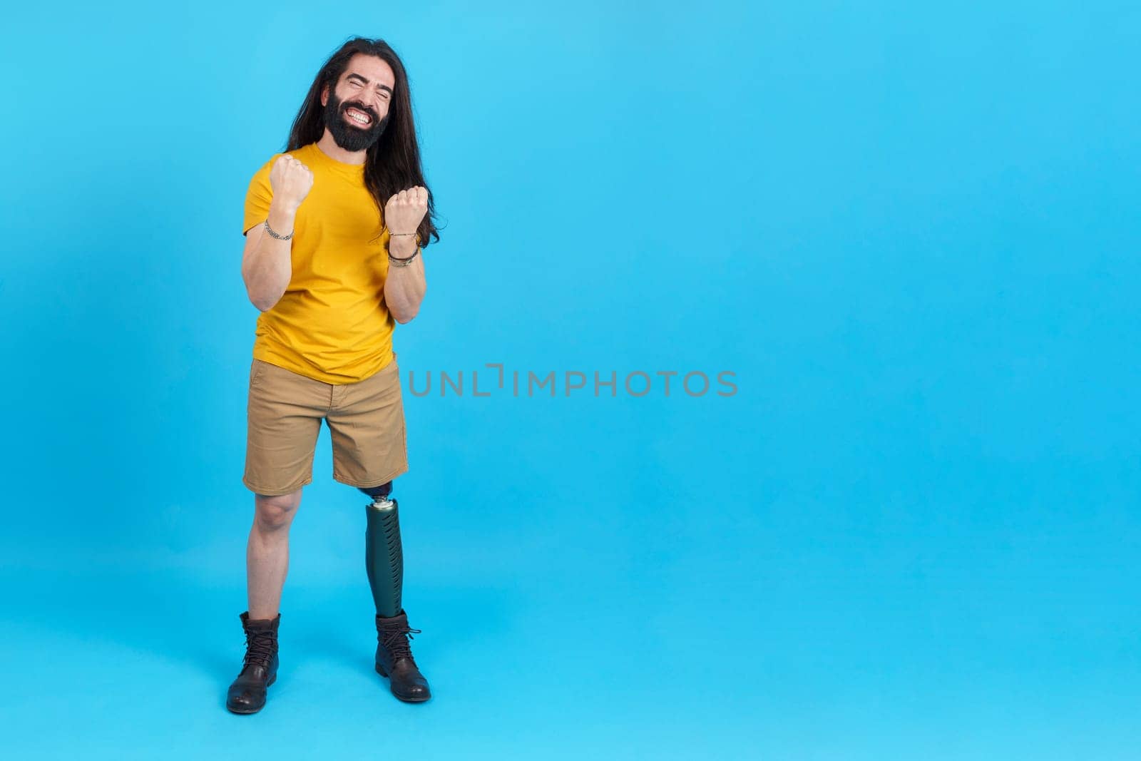 Studio portrait with blue background and copy space of a happy man with a leg prosthesis celebrating with fists in the air