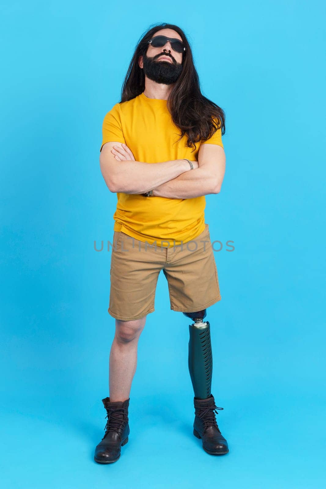 Vertical studio portrait with blue background of a man with long hair and sunglasses standing with a prosthetic leg