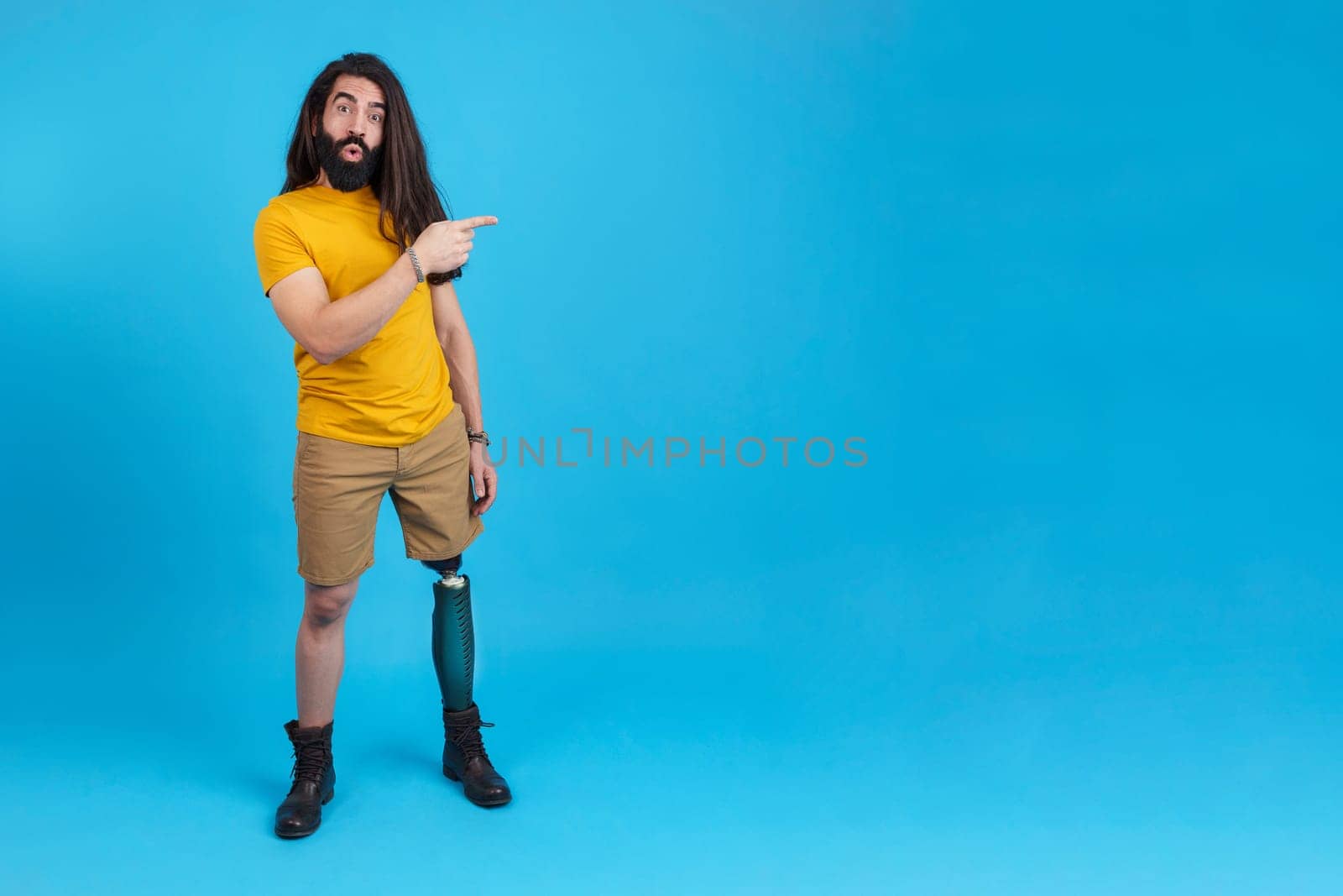 Studio portrait with blue background and copy space of a surprised man with prosthetic leg pointing to the empty space at the side