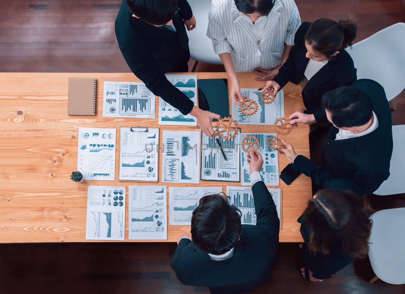Top view hand holding gear by businesspeople wearing suit for harmony synergy in office workplace concept. Group of people hand making chain of gears into collective form with dashboard report papers.