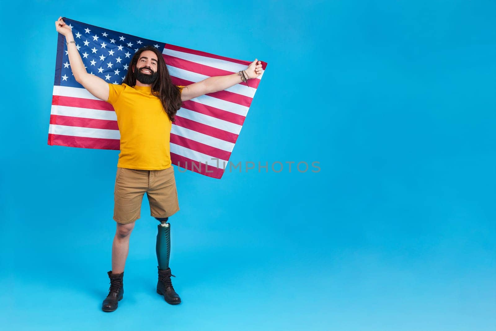 Studio portrait with blue background of a proud man with prosthetic leg raising a north american flag