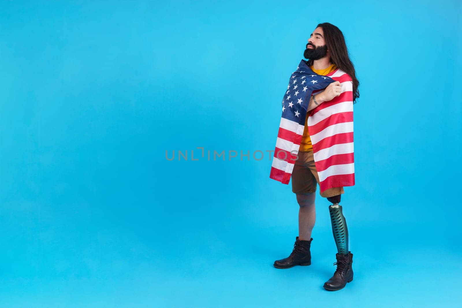 Studio portrait with blue background of a man standing with prosthetic leg wrapped in a north america national flag