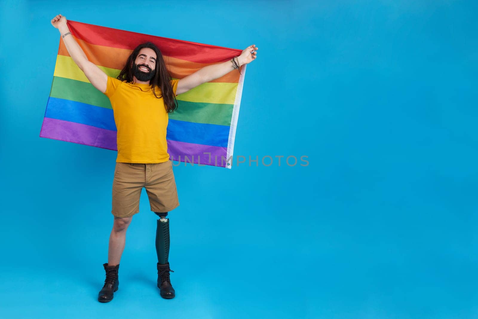 Studio portrait with blue background of a smiley man with a prosthetic leg waving a rainbow lgbt flag