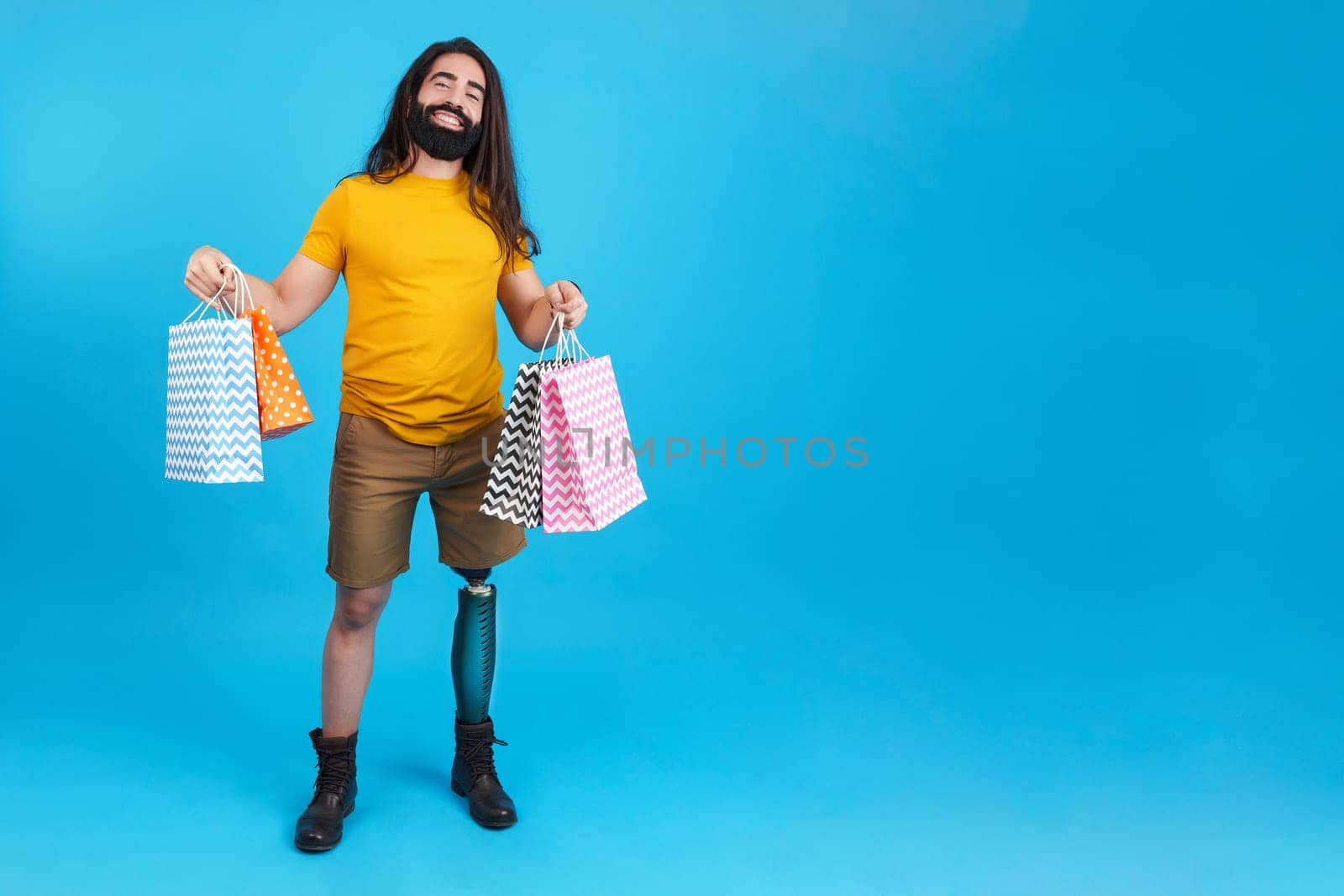 Studio portrait with blue background of a happy man with prosthetic leg holing many shopping bags