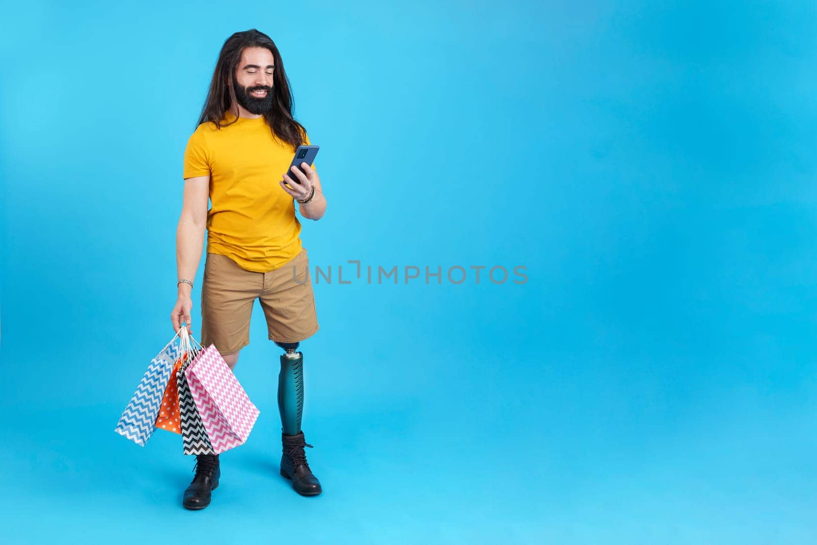 Studio portrait with blue background of a cool man with prosthetic leg using the mobile holing shopping bags