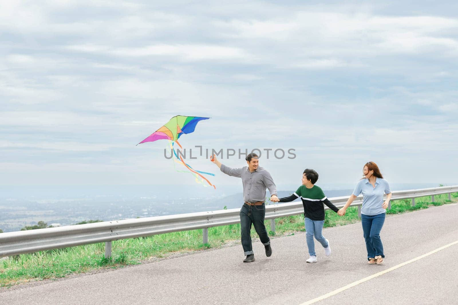 Progressive happy family vacation and carefree day concept. Young parents mother father and son run along and flying kite together road with enjoy natural scenic on scenery and clear sky background.