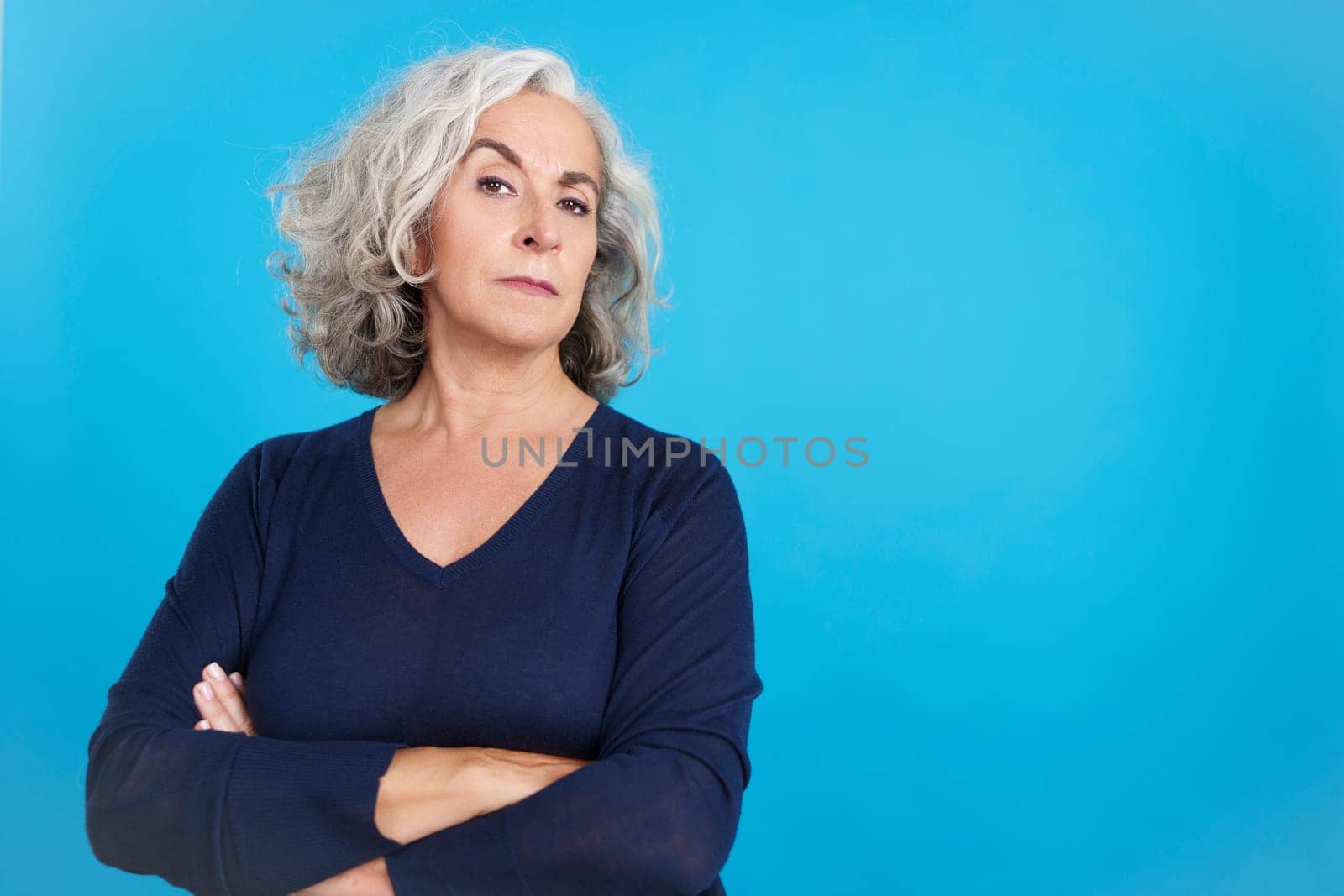Studio portrait with blue background of a mature woman with grey hair looking at camera with a distrustful expression