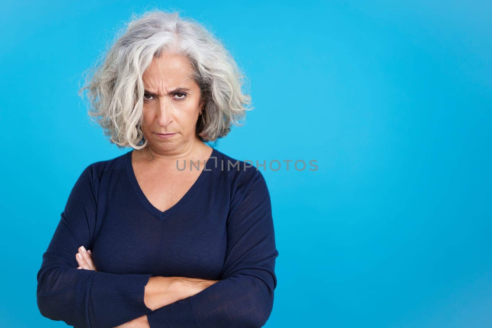 Studio portrait with blue background of a mature woman crossing the arms while looking at camera with an angry expression