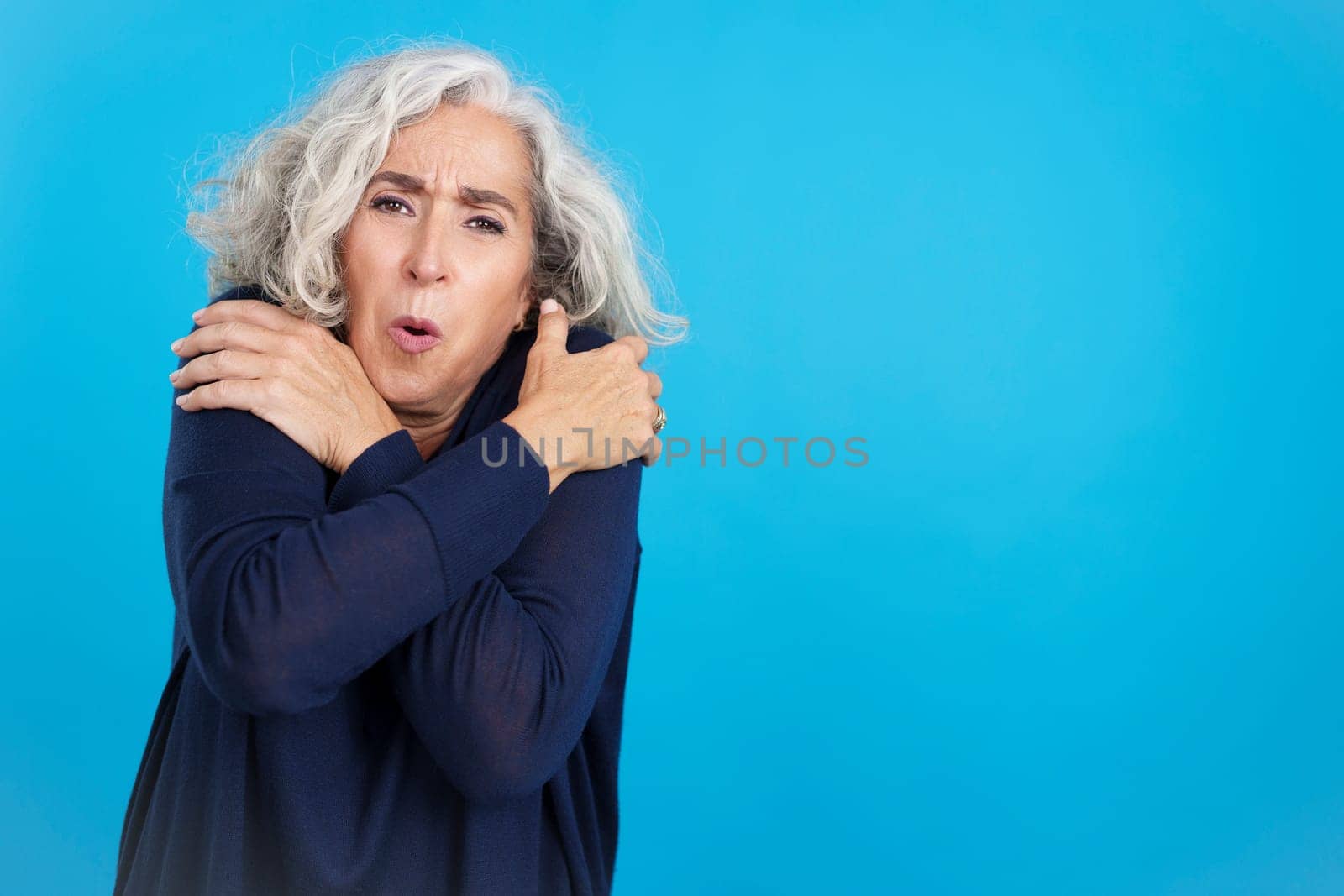 Studio portrait with blue background of an aged caucasian woman looking at camera while embracing herself expressing coldness